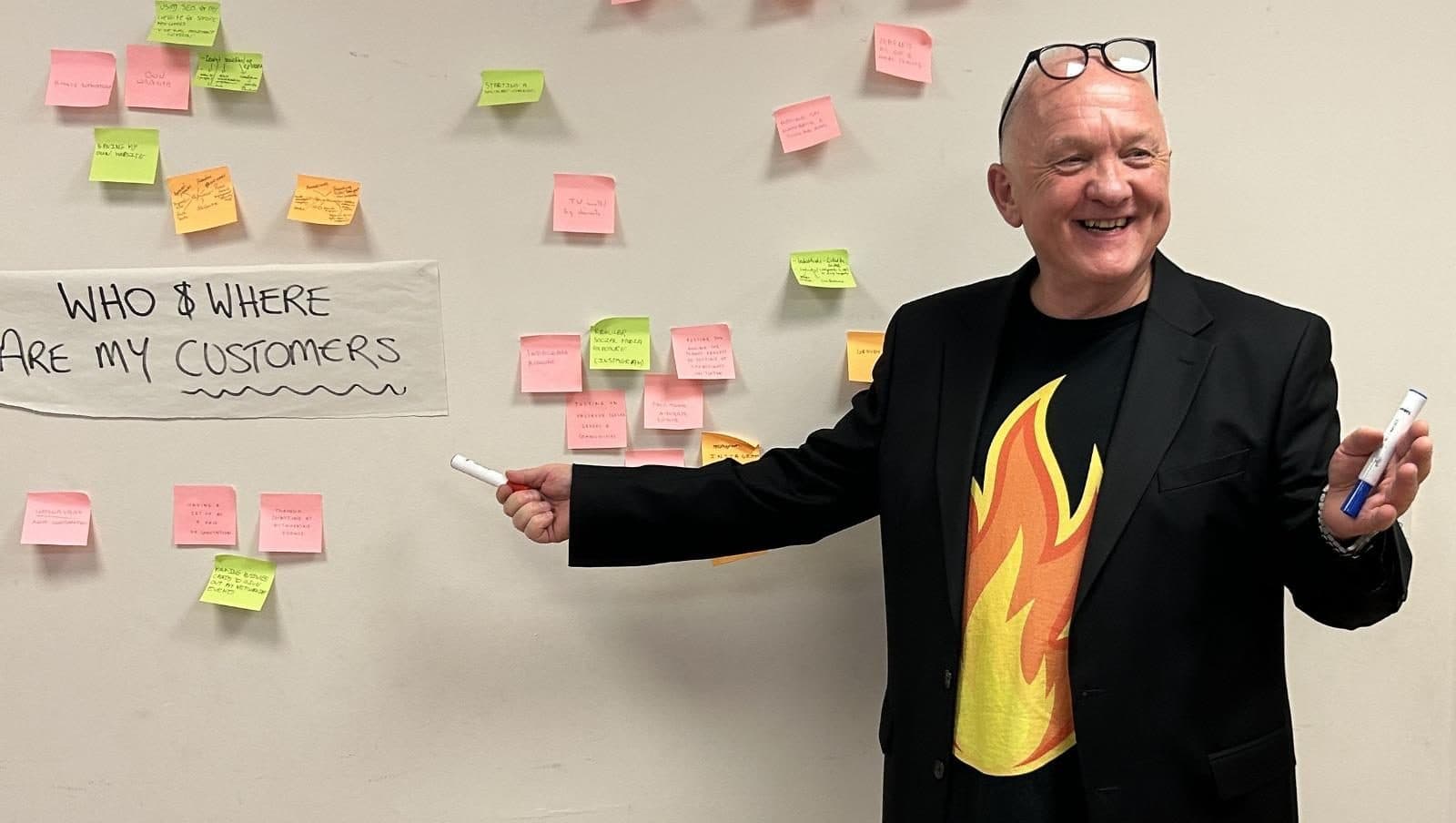 Andrew Bishop smiling beside a wall of sticky notes headed 'Who and where are my customers' at a Firestarter workshop